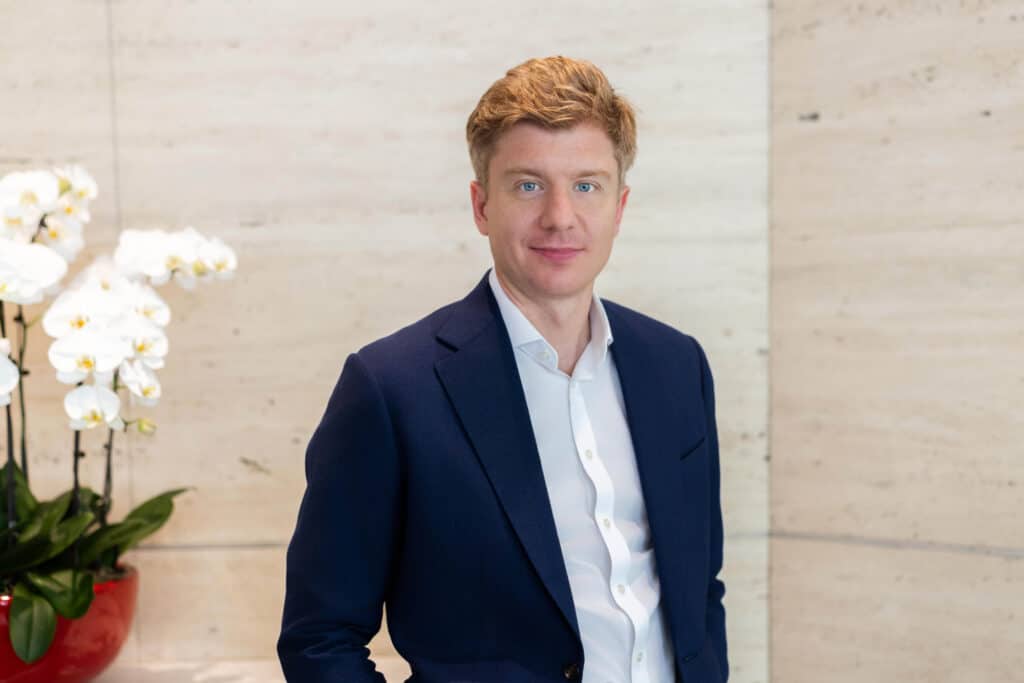 Portrait of a man with reddish-blonde hair and blue eyes, wearing a blue suit jacket and white shirt, standing in front of a light-colored wall with white orchids.