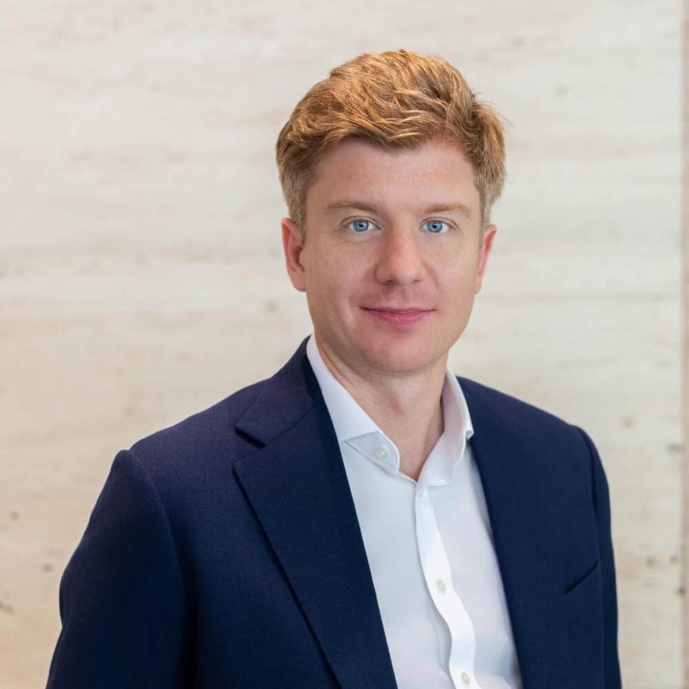 Portrait of a man with reddish-blonde hair and blue eyes, wearing a blue suit jacket and white shirt, standing in front of a light-colored wall with white orchids.