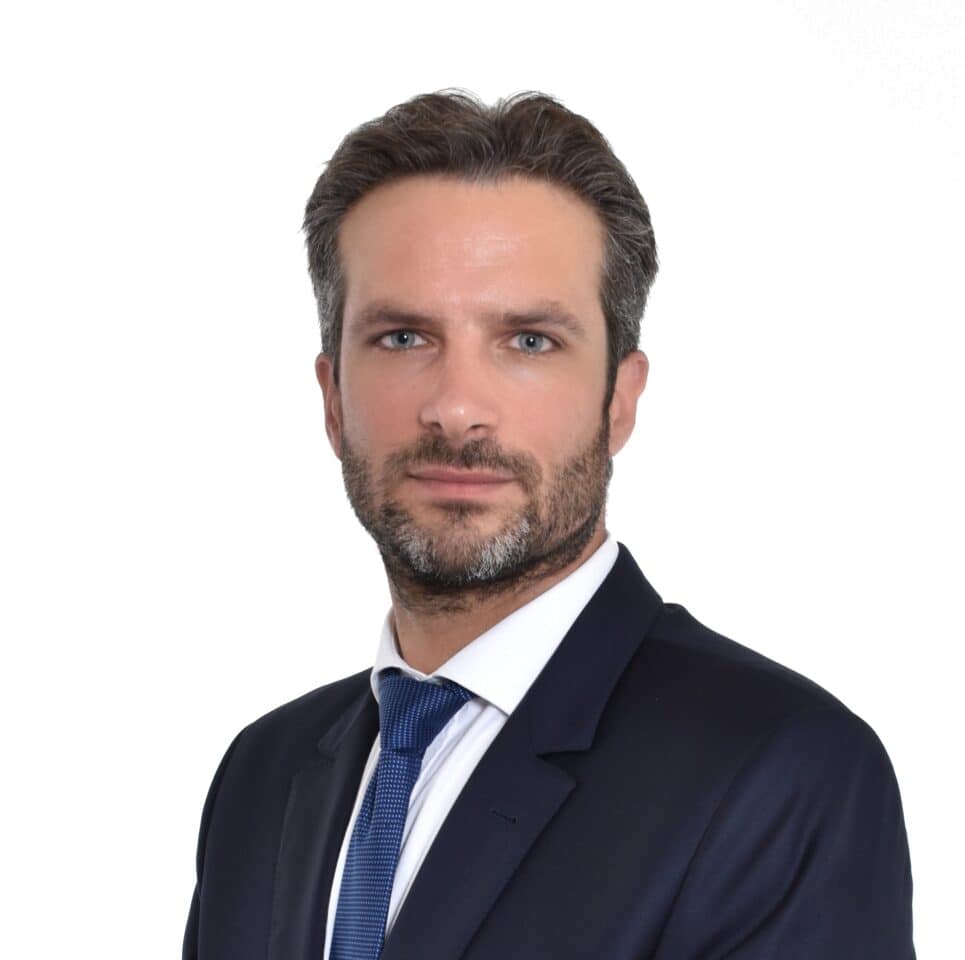 Headshot of a man with dark hair and beard, wearing a dark suit and blue tie against a white background.