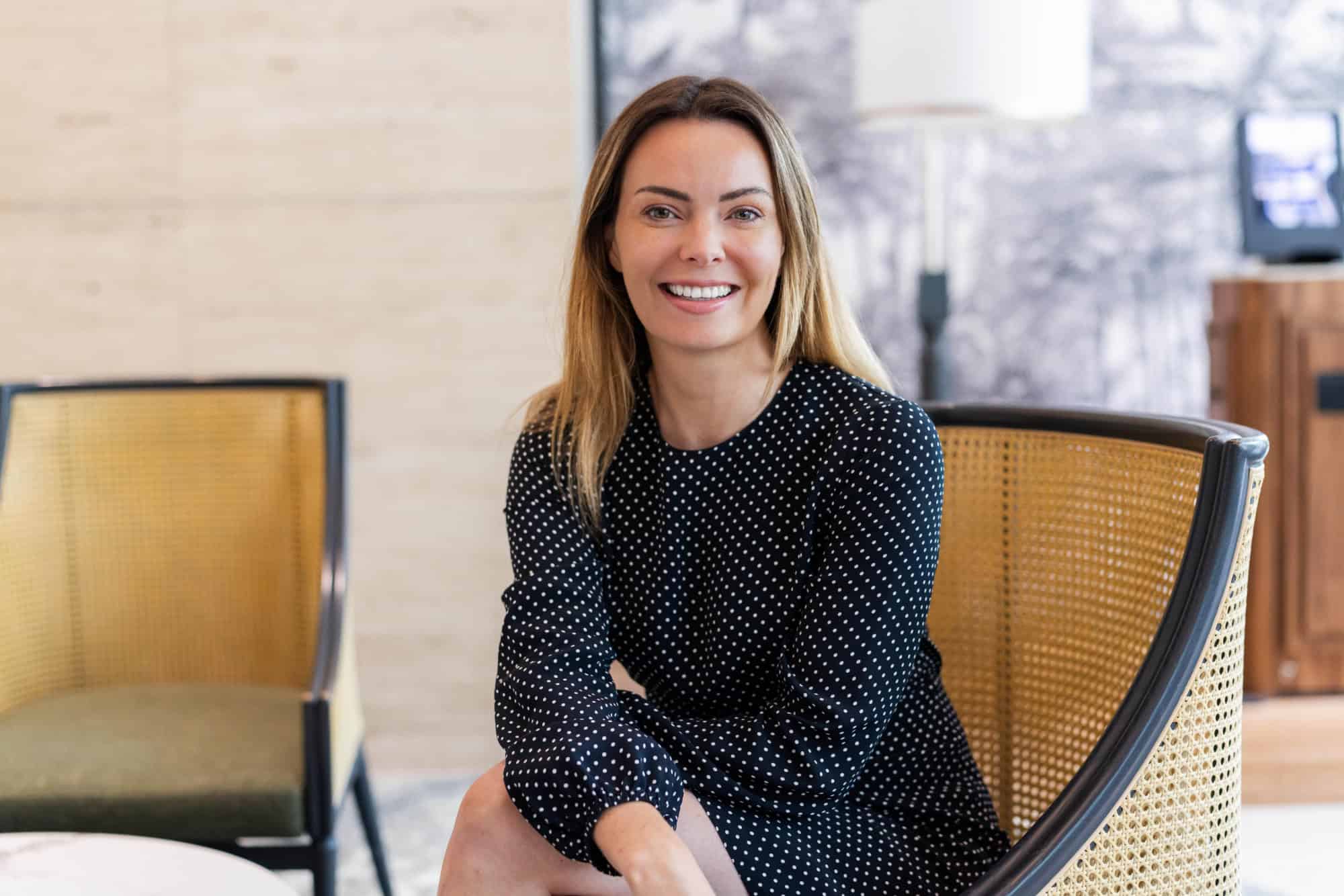 A woman with long hair and a black polka dot dress smiles while sitting in a cane armchair.
