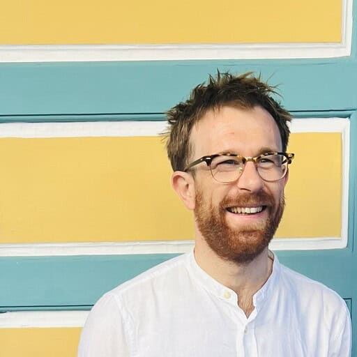 A smiling man with a beard and glasses in a white shirt, in front of a yellow and blue striped wall.