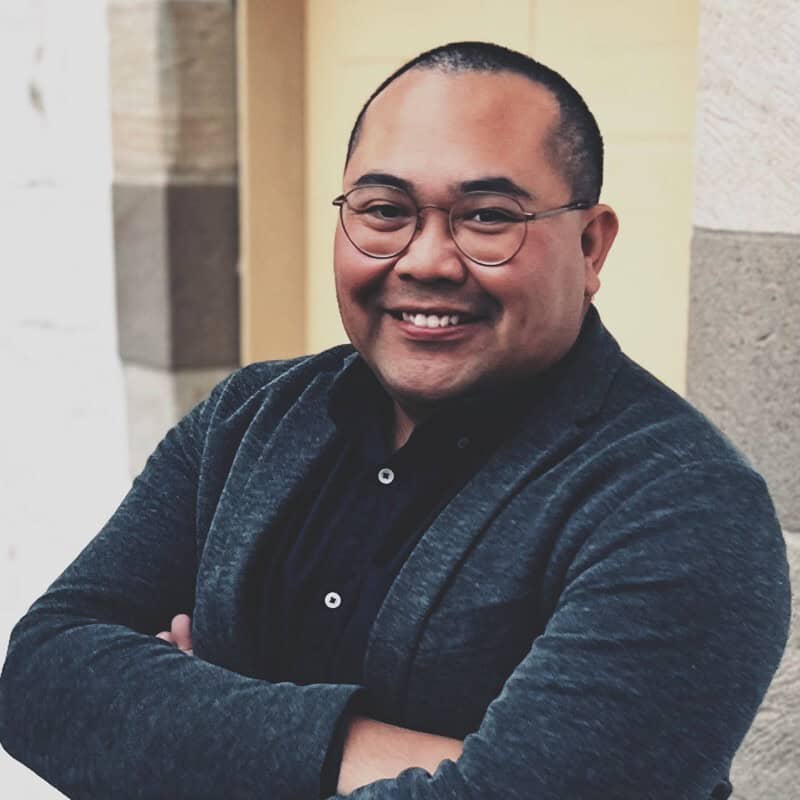 A smiling man with short hair and round glasses, wearing a dark shirt and gray blazer, with arms crossed.