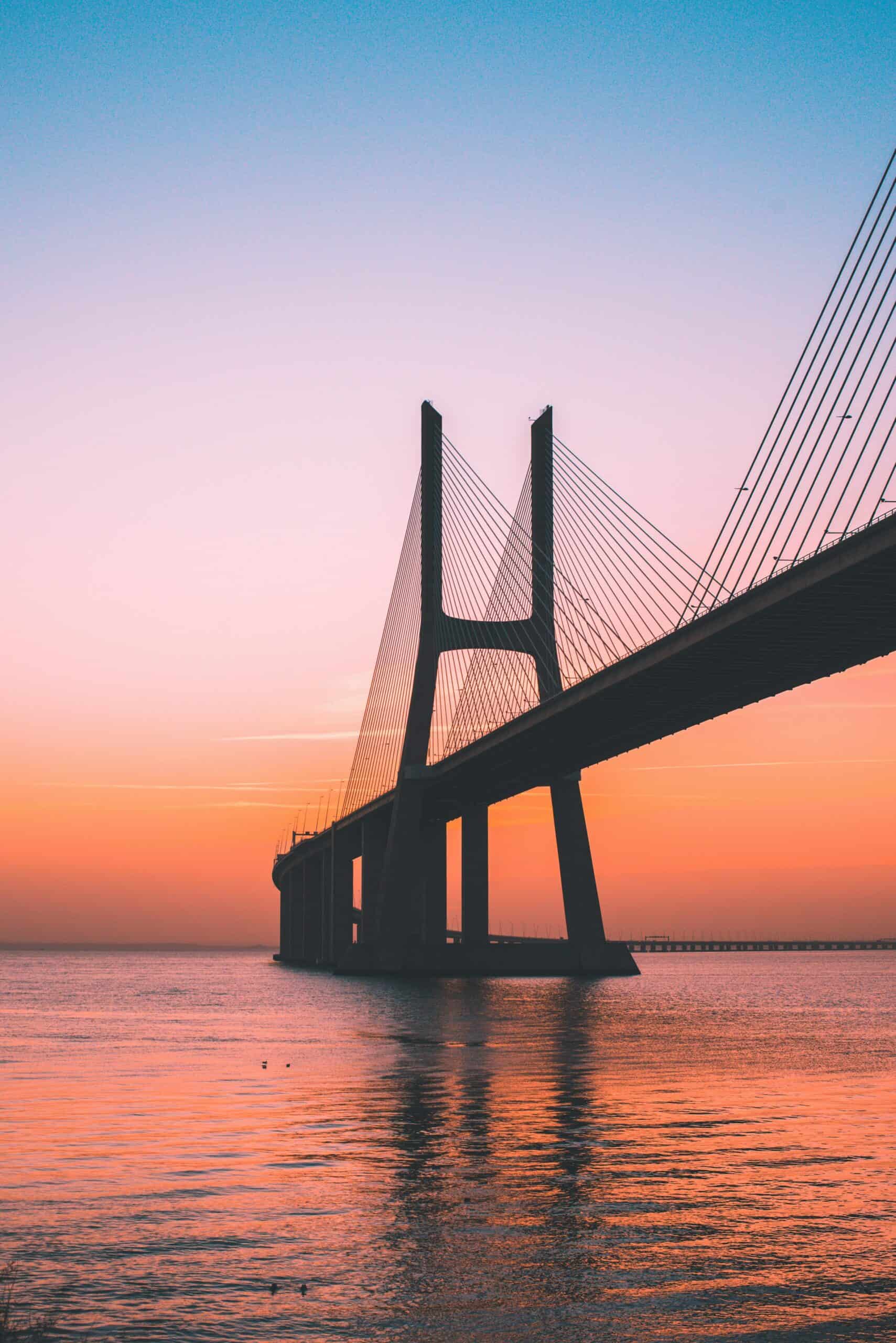 Silhouette of the Vasco da Gama Bridge over water at sunset, with a sky transitioning from blue to pink to orange.