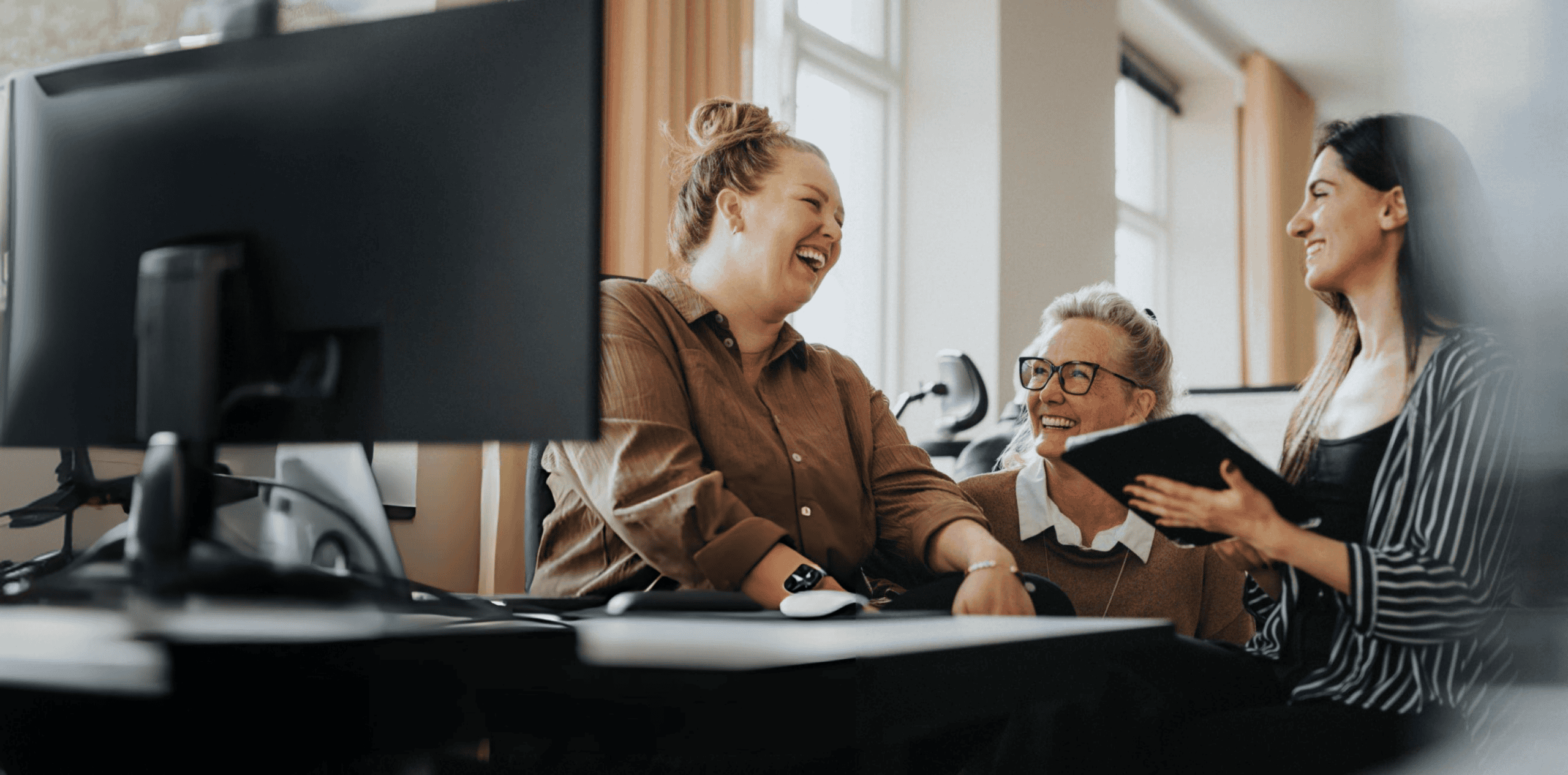 Three women smiling and laughing together in an office, one holding a tablet, with a computer monitor in the foreground.