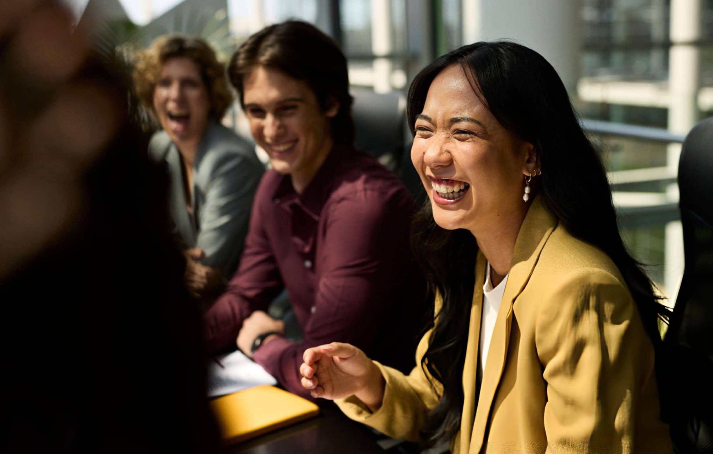 Three people laughing in a meeting room. An Asian woman in a yellow blazer laughs heartily in the foreground, with a man and another woman smiling and laughing behind her.