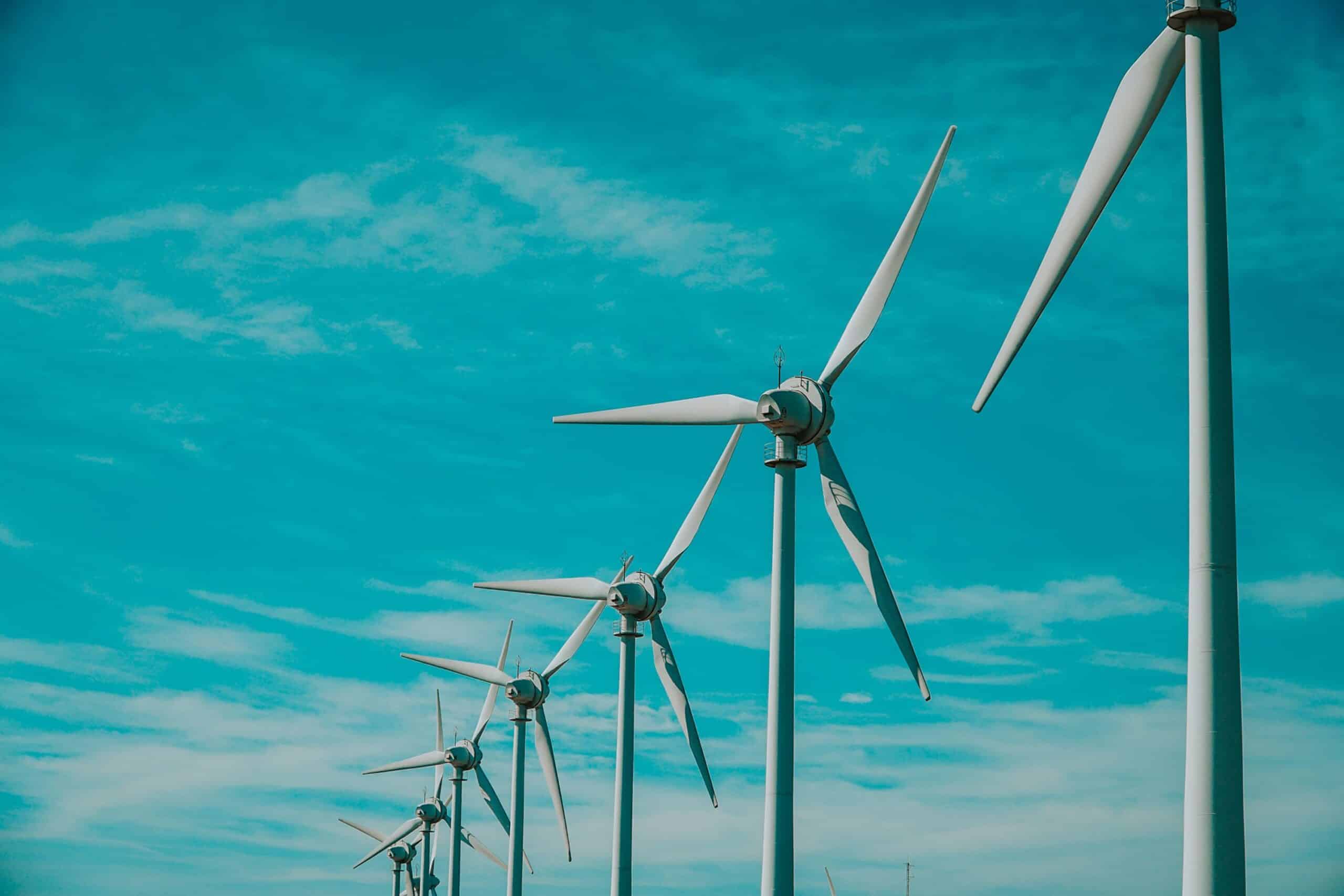 A row of white wind turbines with three blades each, standing against a bright turquoise sky with scattered white clouds.