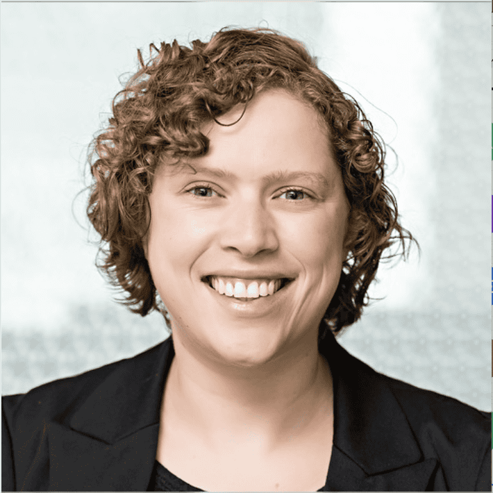 Headshot of a smiling woman with curly brown hair and blue eyes, wearing a black blazer.