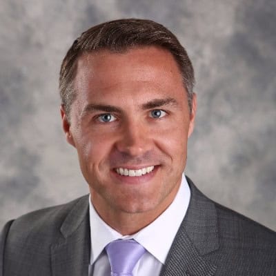 Headshot of a smiling man with short brown hair, blue eyes, wearing a gray suit and a lavender tie.