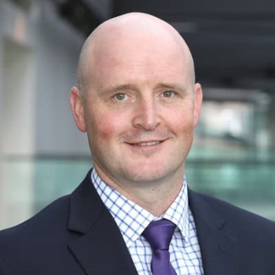 Headshot of a bald man smiling, wearing a dark blue suit, a blue and white checkered shirt, and a purple tie.