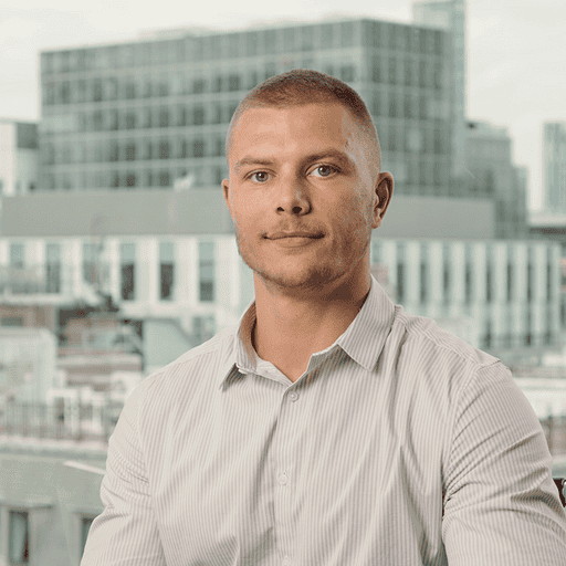 Headshot of a man with short hair and a striped shirt, looking forward, with blurred city buildings in the background.