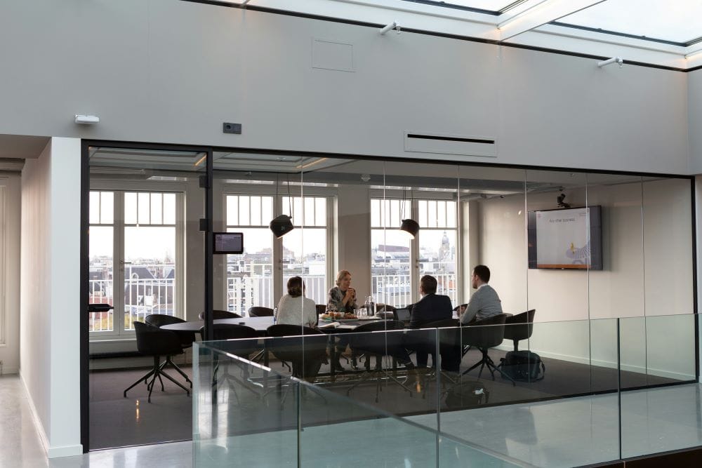 Four people in a glass-walled conference room with a city view, seated around a table with a presentation on a screen.