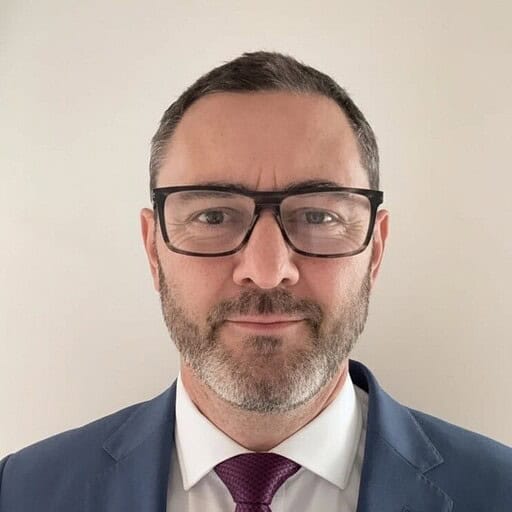 Headshot of a man with short dark hair, beard, glasses, blue suit, and purple tie.