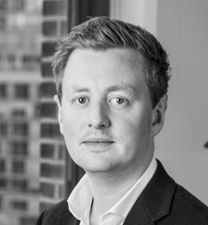 Black and white portrait of a man with short hair, wearing a suit jacket and open-collared shirt, looking forward.