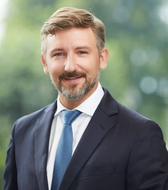 Headshot of a smiling man with a beard, blue eyes, and light brown hair, wearing a dark suit and blue tie against a blurred green background.
