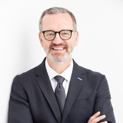 Headshot of a smiling man with gray hair and beard, wearing glasses, a dark suit, white shirt, and dark tie, against a white background.