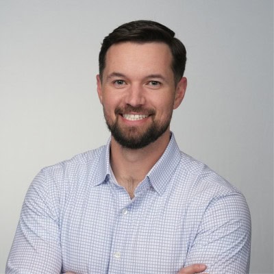 Headshot of a smiling man with dark hair and a beard, wearing a light blue checkered shirt.