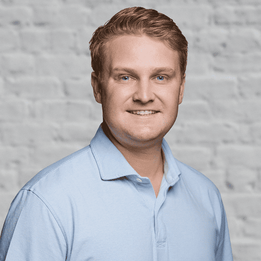 Headshot of a smiling man with light red hair and blue eyes, wearing a light blue polo shirt, against a gray brick wall.