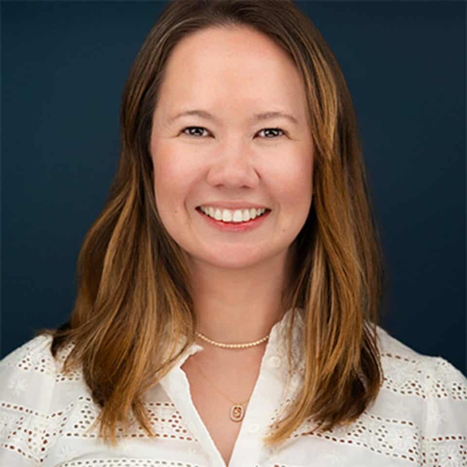 Headshot of a smiling woman with long brown hair, wearing a white embroidered shirt and gold necklaces, against a dark blue background.