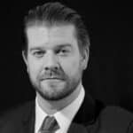 Black and white portrait of a man with a beard and swept-back hair, wearing a suit and tie.