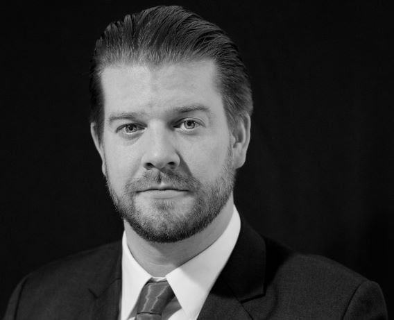 Black and white portrait of a man with a beard and swept-back hair, wearing a suit and tie.