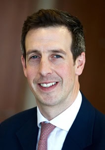Headshot of a smiling man with short dark hair, wearing a dark suit and red patterned tie.
