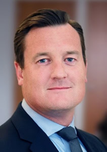 Headshot of a man with dark hair and blue eyes, wearing a dark suit and light blue shirt.