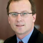 Headshot of a man with short brown hair and glasses, wearing a suit and tie.