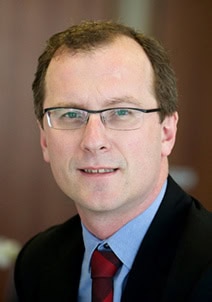 Headshot of a man with short brown hair and glasses, wearing a suit and tie.