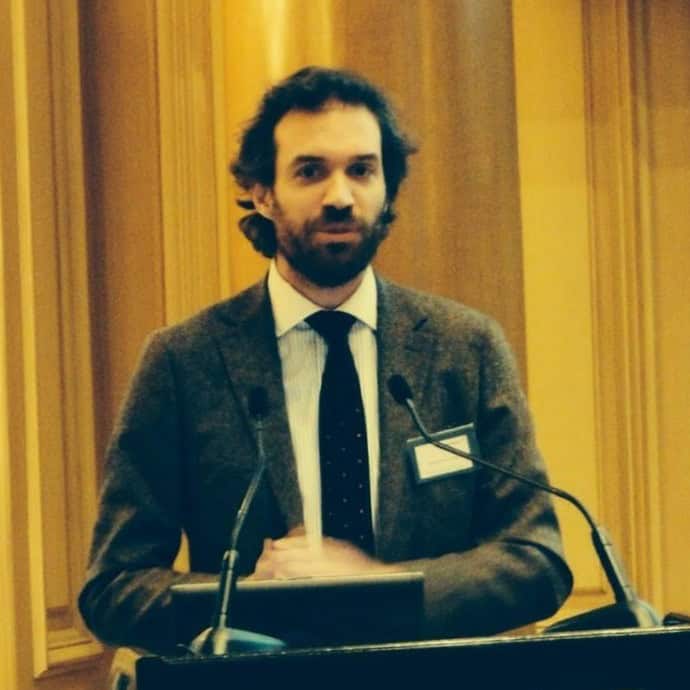 A man with dark hair and a beard in a suit jacket speaks at a podium with microphones.
