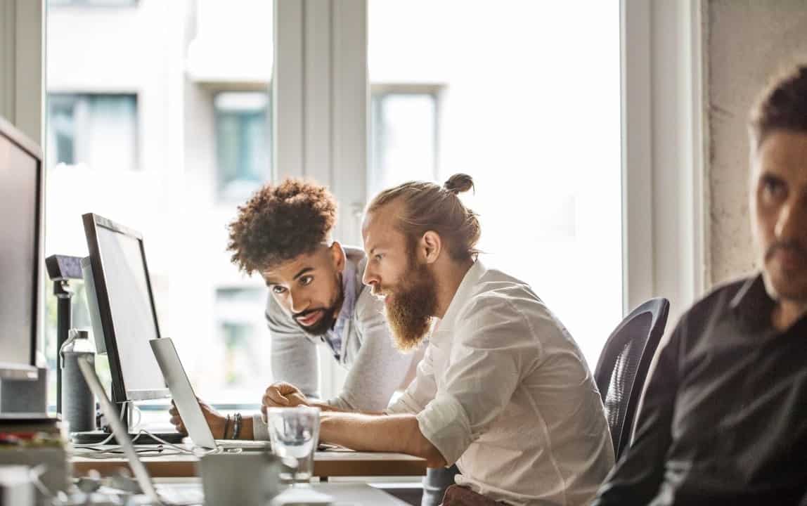 Two men, one with a beard and hair in a bun, and another with curly hair, lean over a laptop at a desk in a bright office, looking at the screen together.