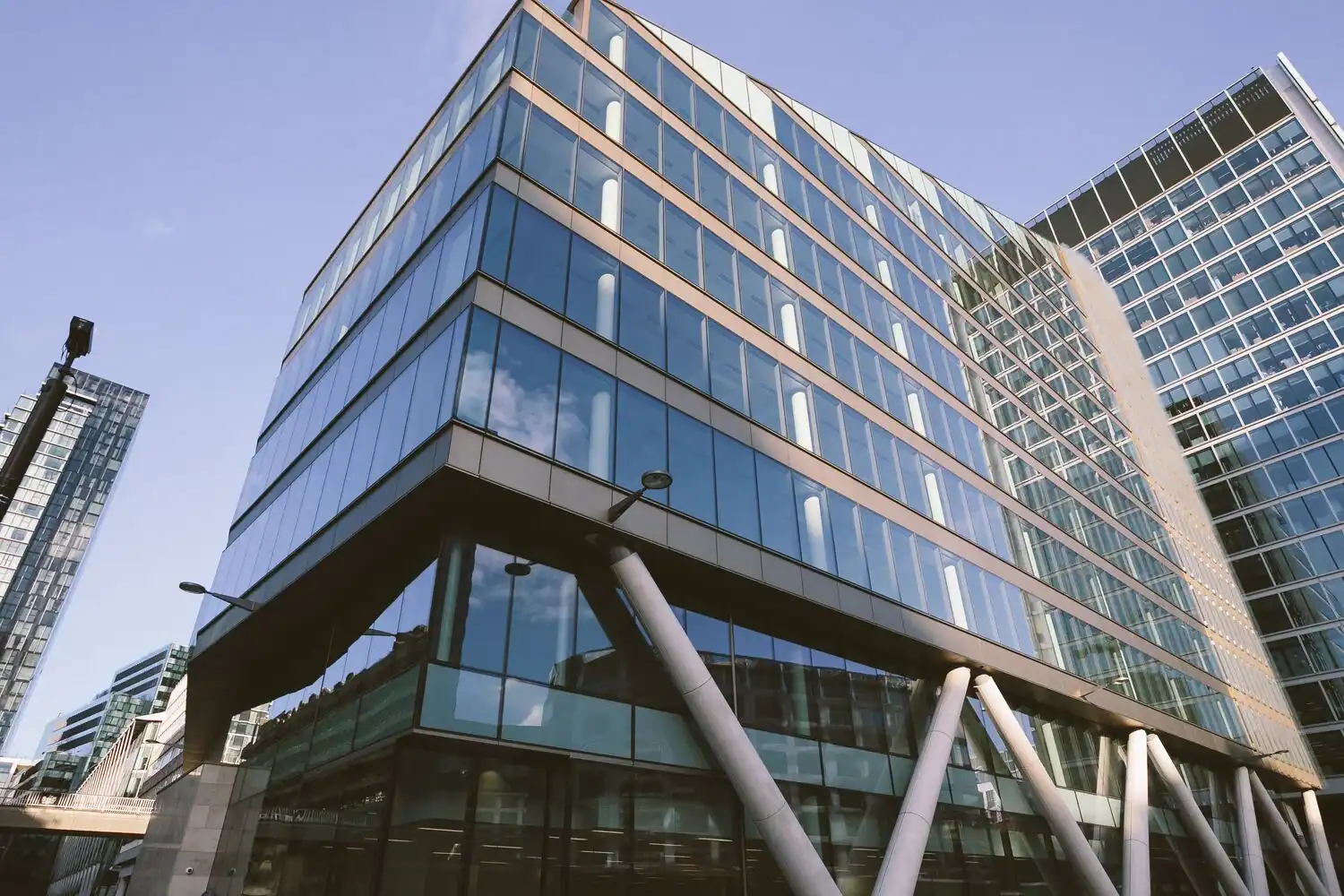 Low-angle view of a modern glass-clad building with angled white support columns, reflecting a blue sky and clouds.