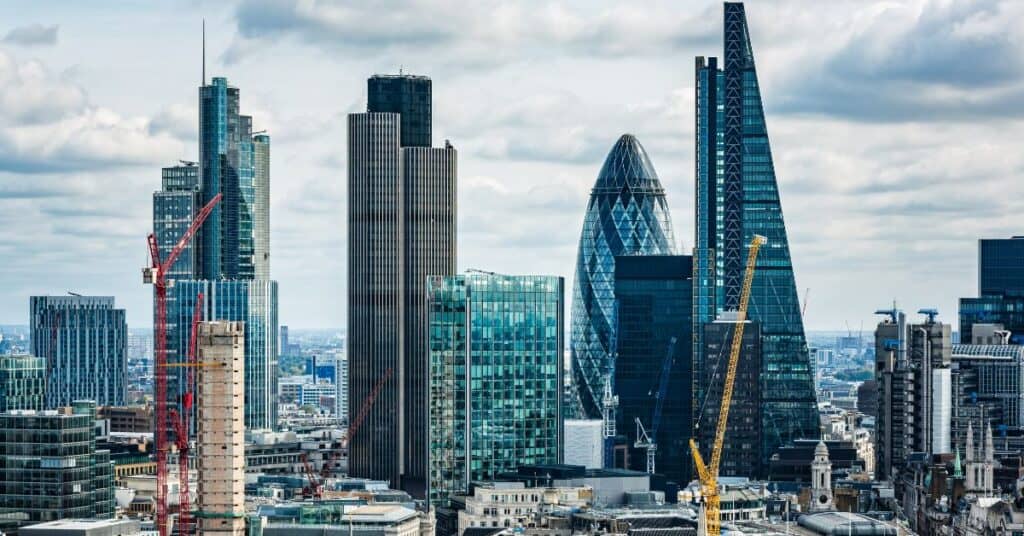 A city skyline featuring numerous modern skyscrapers, including The Gherkin, and several construction cranes under a cloudy sky.