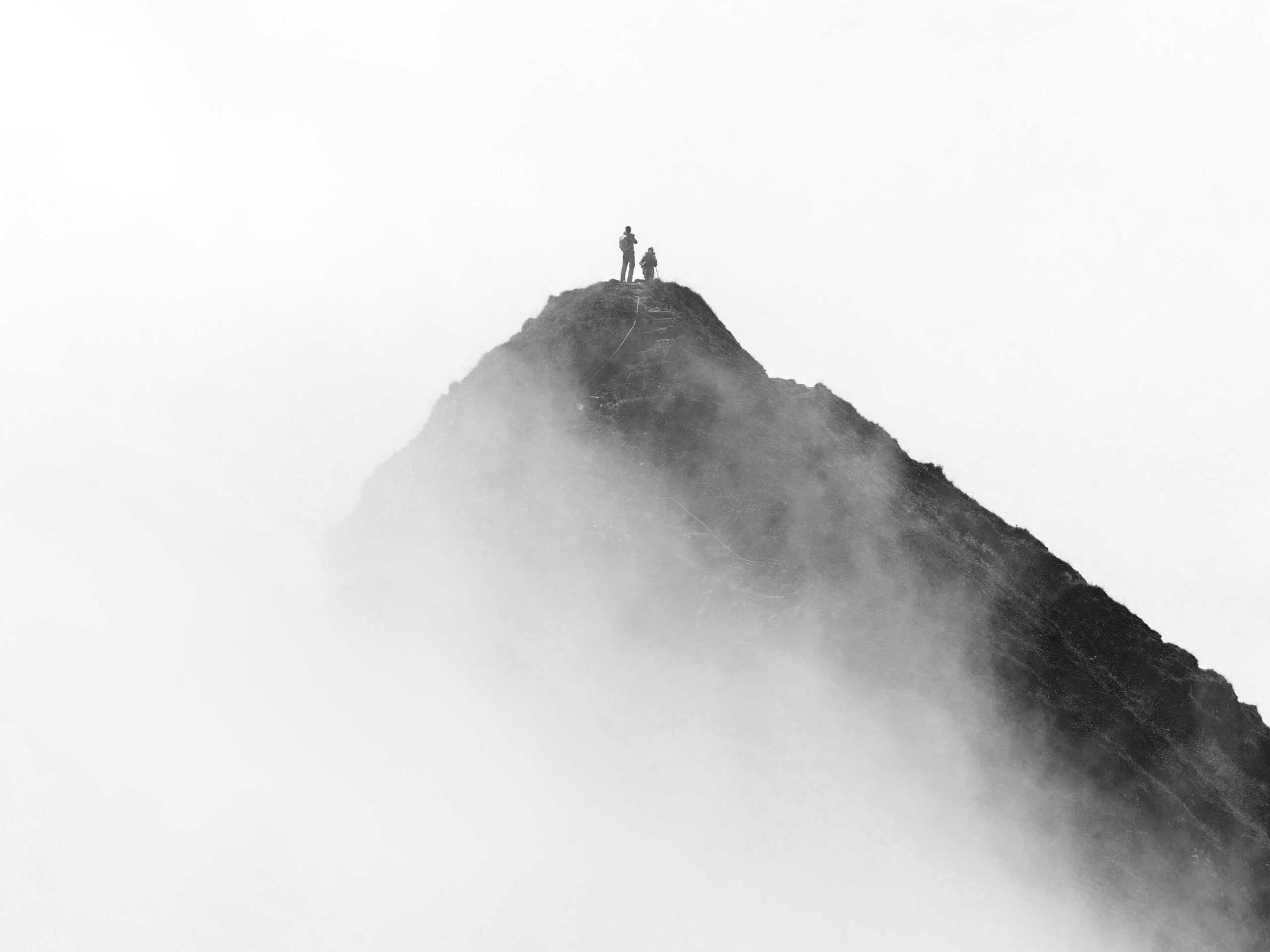 Black and white image of two small figures on a foggy mountain summit, with a rope visible on the slope.