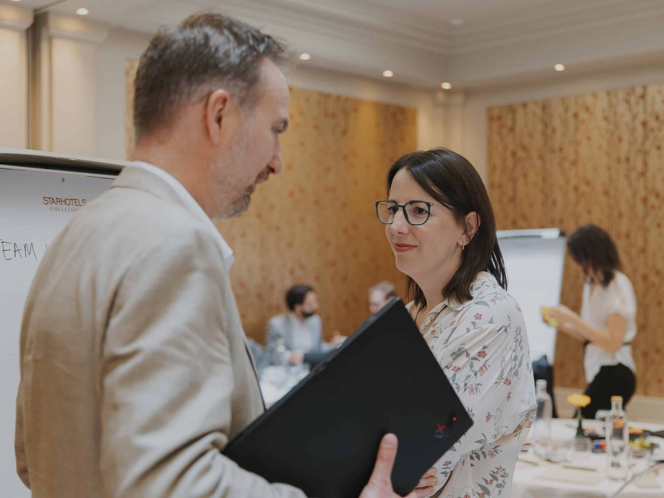 A man and a woman conversing in a meeting room. The man is seen from behind, holding a laptop, while the woman faces him, smiling and wearing glasses and a floral blouse. A Starhotels flipchart is visible in the background.