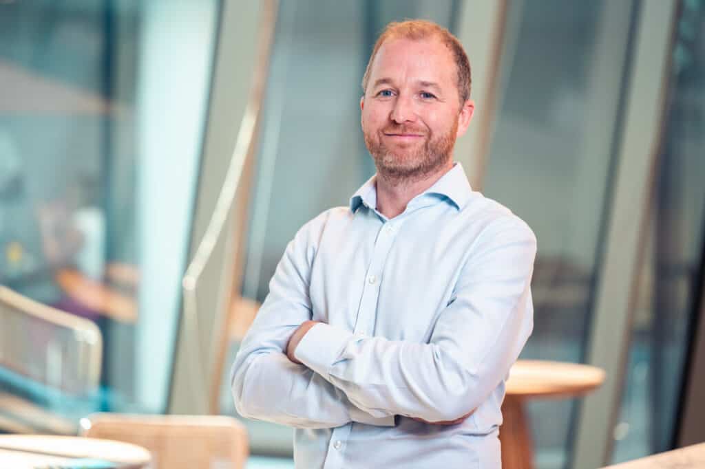A man with a beard and light blue shirt smiles with arms crossed in a modern office.