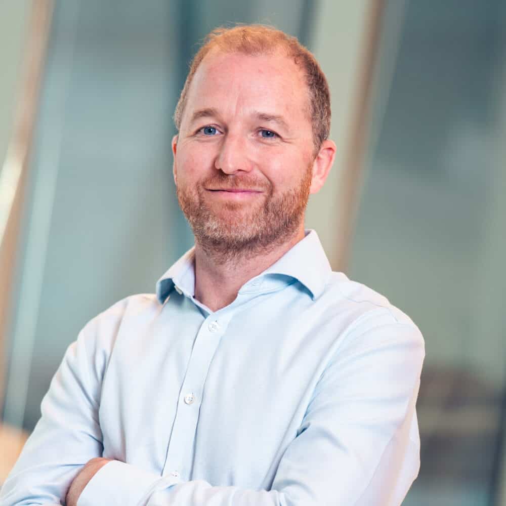 A man with a beard and light blue shirt smiles with arms crossed in a modern office.