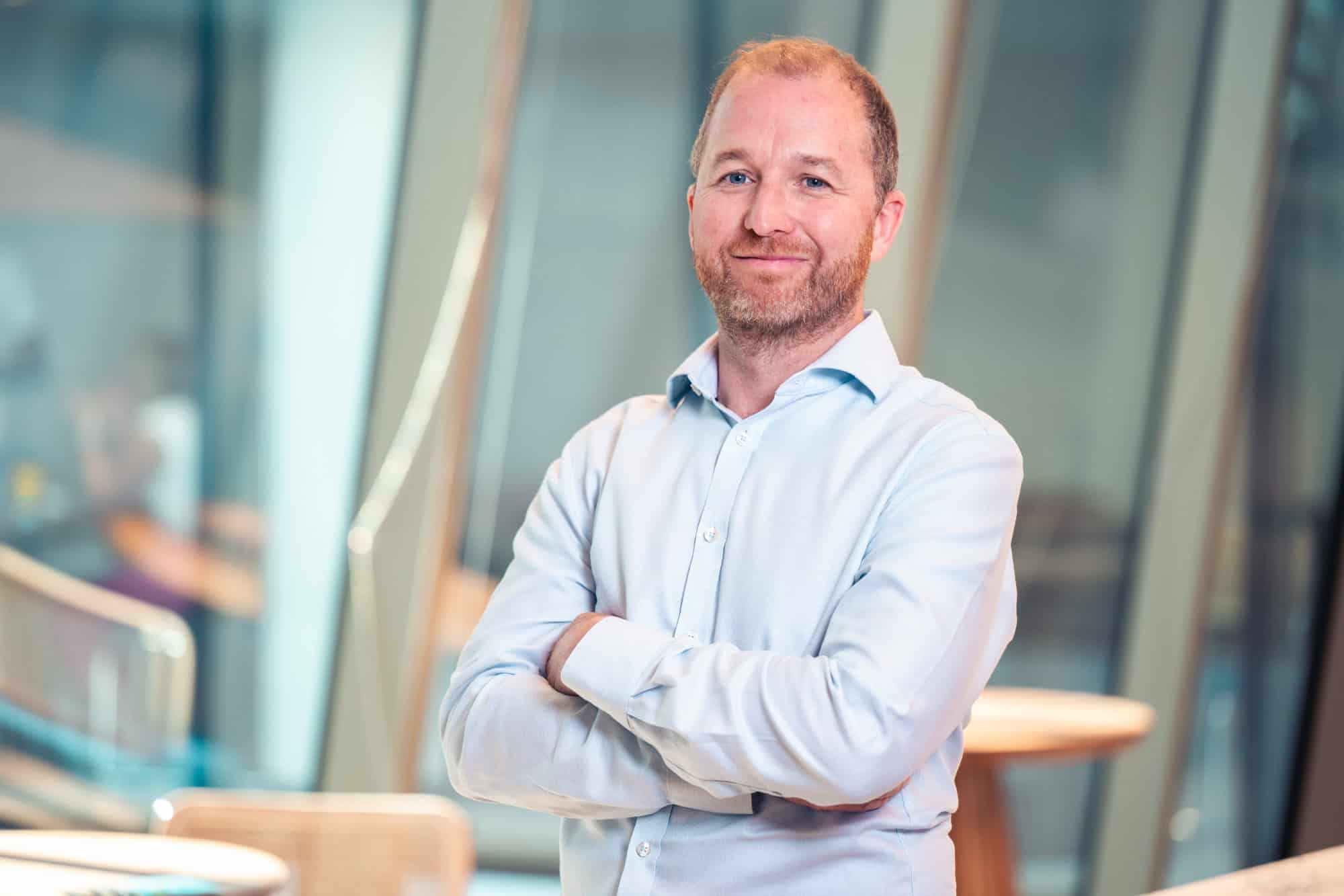 A man with a beard and light blue shirt smiles with arms crossed in a modern office.