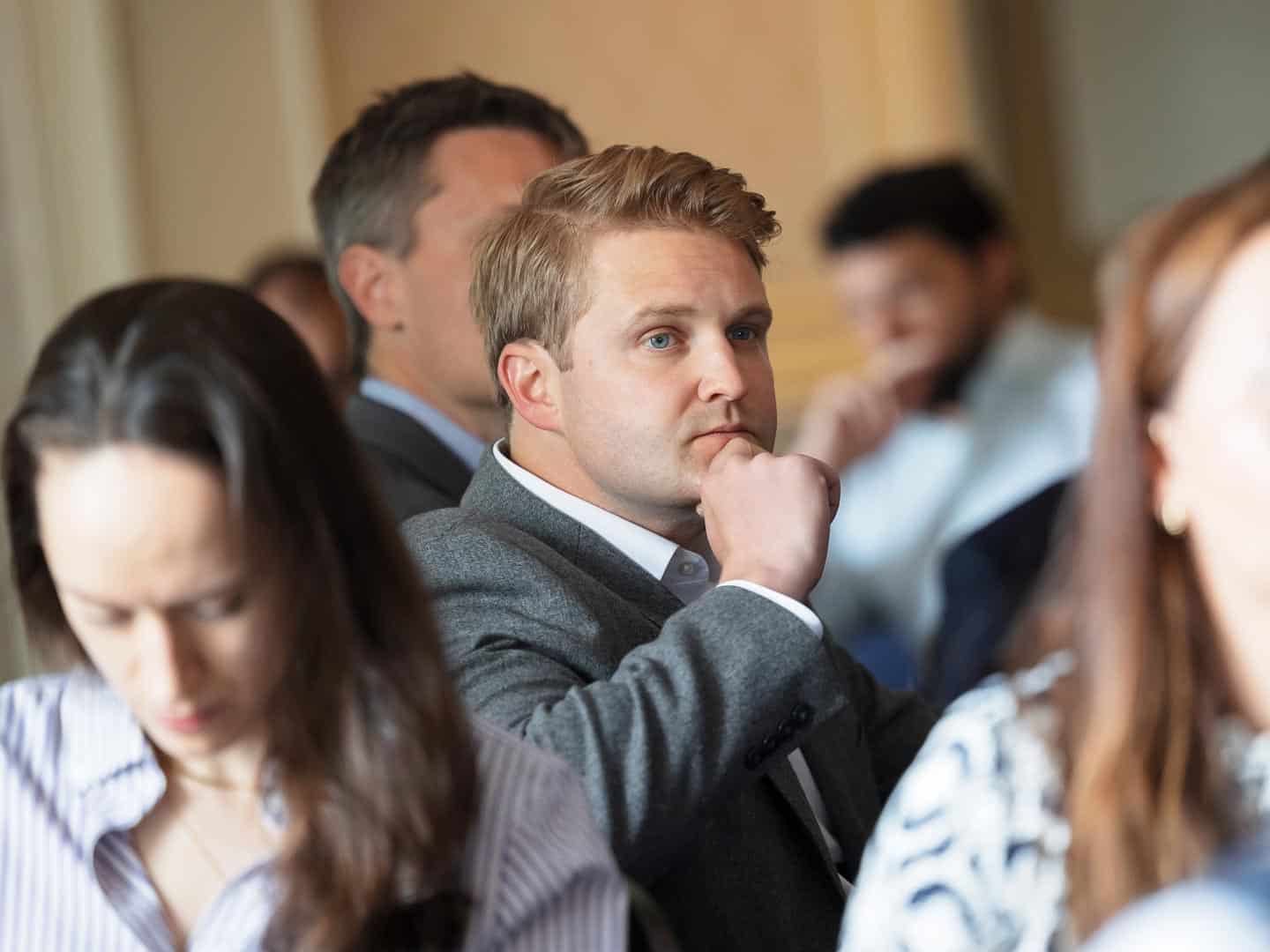 A man with blonde hair and blue eyes in a grey jacket, hand to chin, looking thoughtfully to the side at an event.