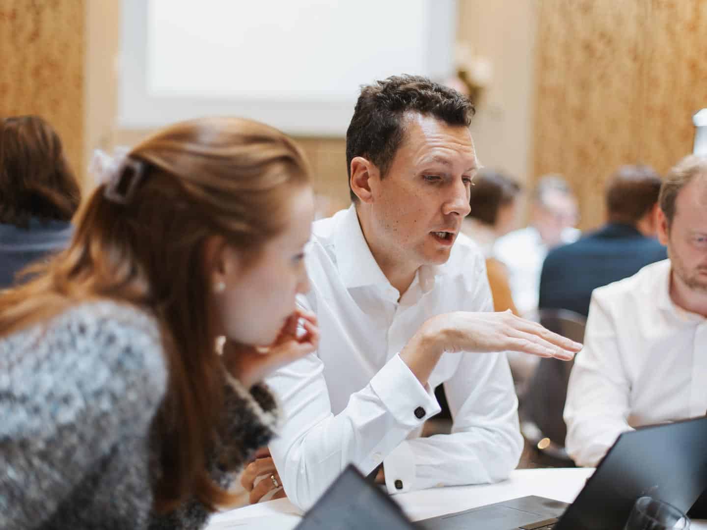 A man in a white shirt gestures while speaking to a woman with reddish-brown hair who is listening intently. Laptops are on the table, and other people are blurred in the background.