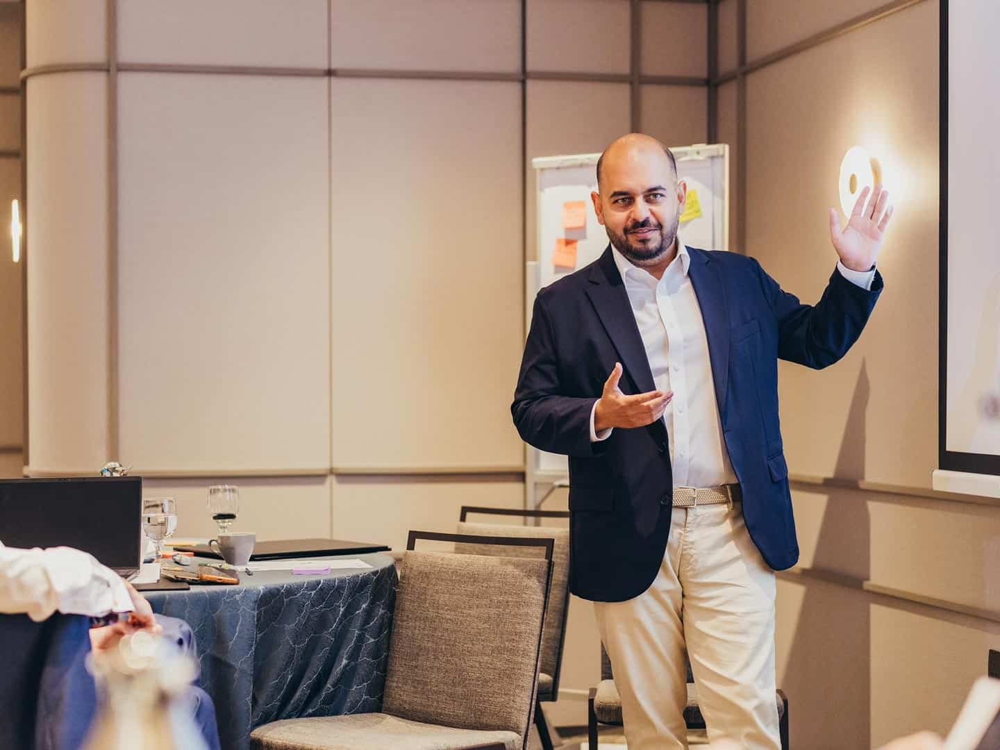 A man with a beard in a blazer and khaki pants presents in a conference room, gesturing with both hands.