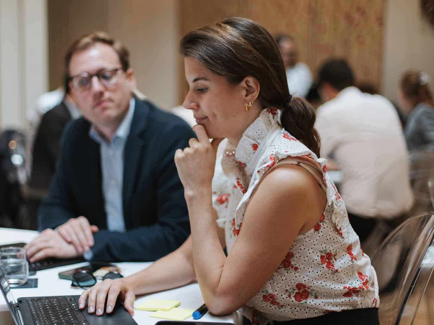 A woman with dark hair in a floral top works on a laptop, with a blurred man in glasses beside her at a table.