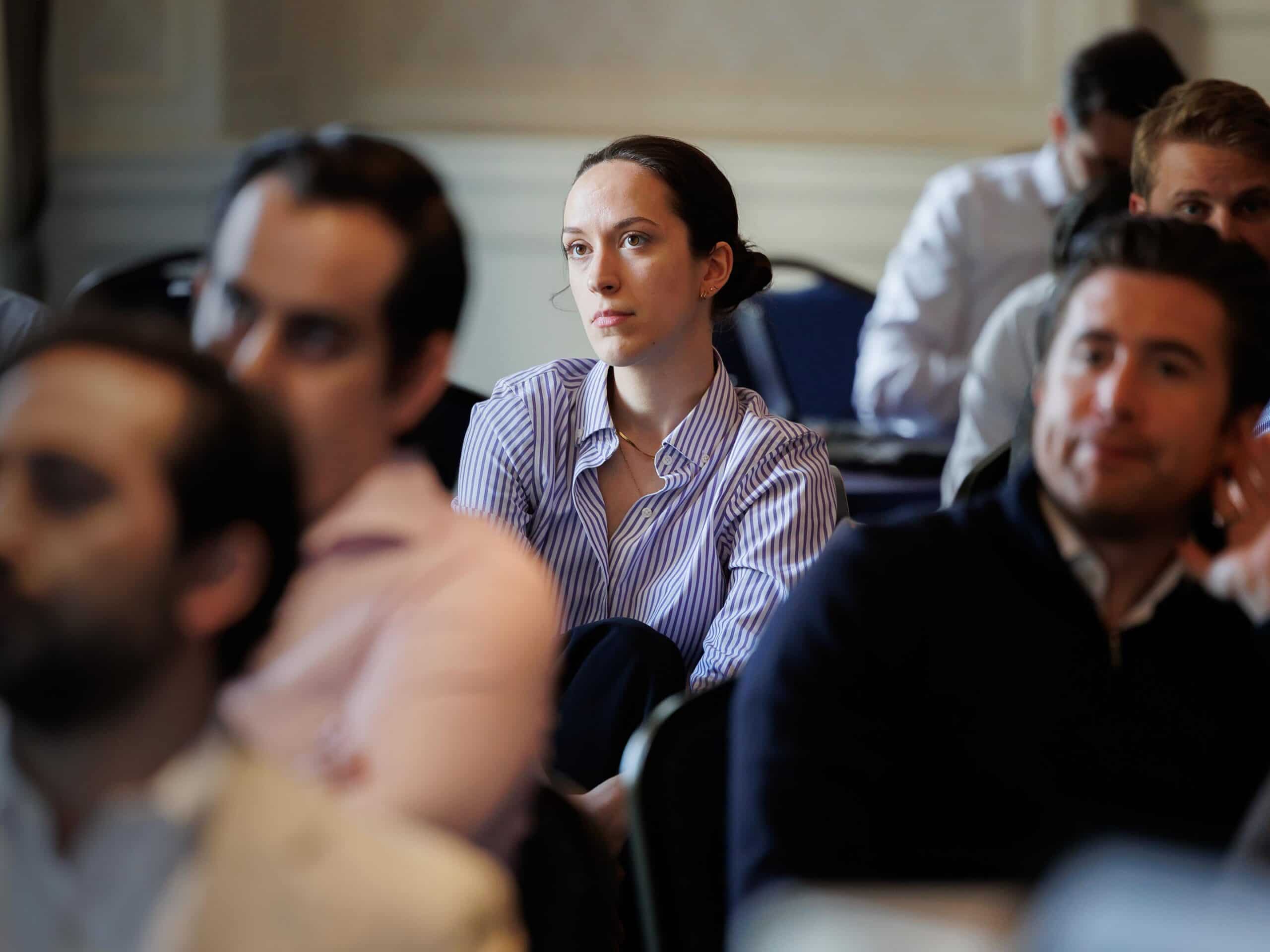 A woman with dark hair pulled back, wearing a blue and white striped shirt, looks attentively to the side while seated among other people at an event.