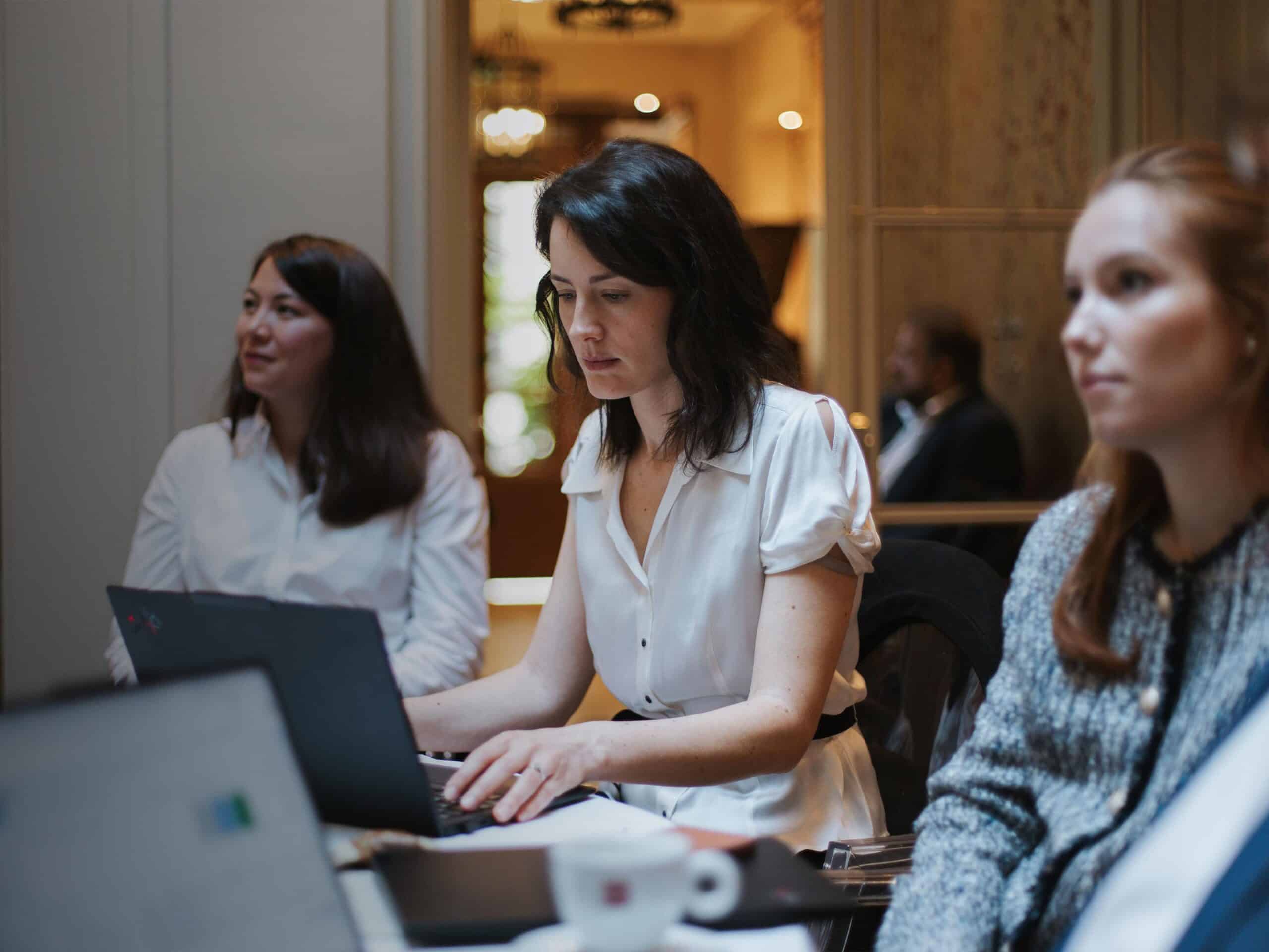 Three women in a meeting, one typing on a laptop, with two others partially visible.