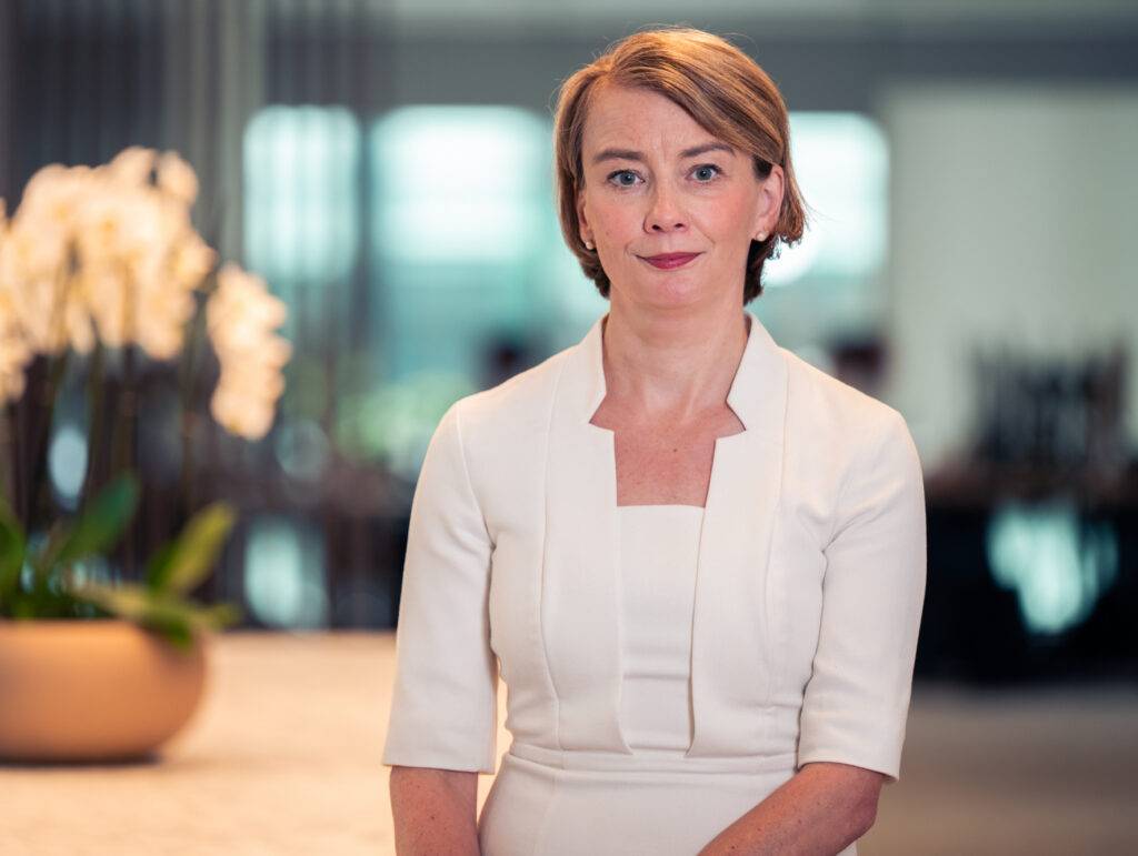 A woman with short brown hair and blue eyes, wearing a cream dress, looks directly at the camera with a slight smile. A blurred orchid plant is in the background.