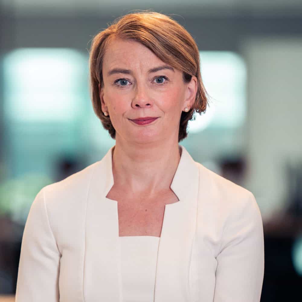 A woman with short brown hair and blue eyes, wearing a cream dress, looks directly at the camera with a slight smile. A blurred orchid plant is in the background.