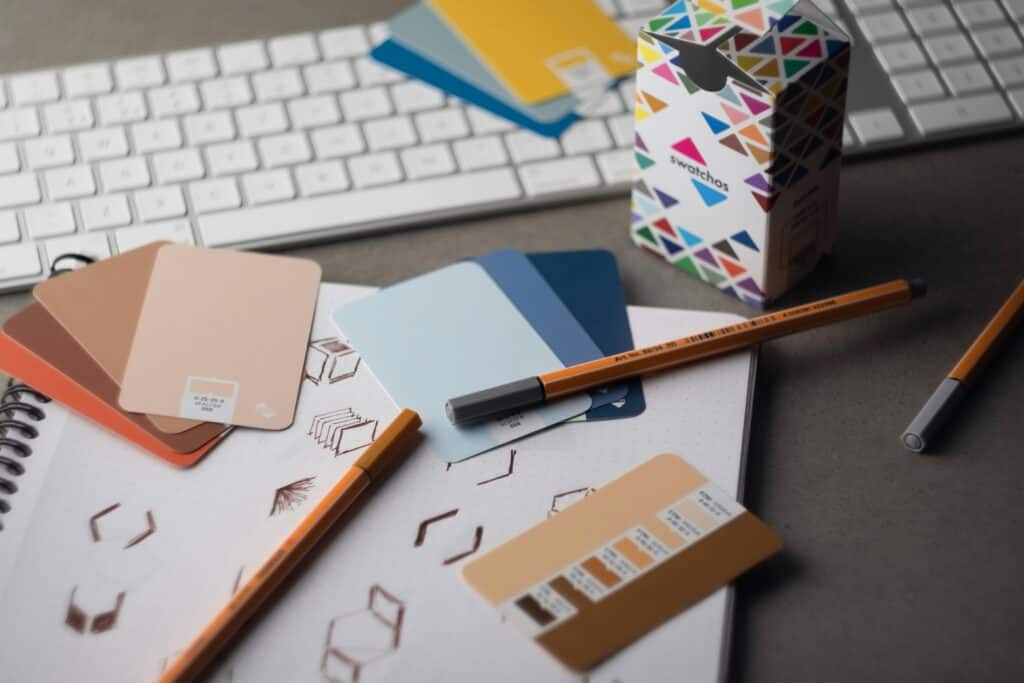 A flat lay of design tools including a white keyboard, a notebook with sketches, various color swatch cards in peach, blue, and brown tones, two orange pens, and a 'swatchos' box on a dark gray surface.