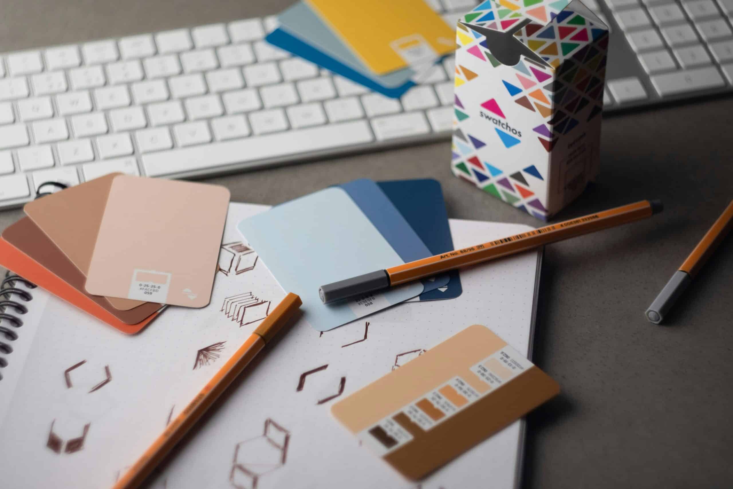 A flat lay of design tools including a white keyboard, a notebook with sketches, various color swatch cards in peach, blue, and brown tones, two orange pens, and a 'swatchos' box on a dark gray surface.