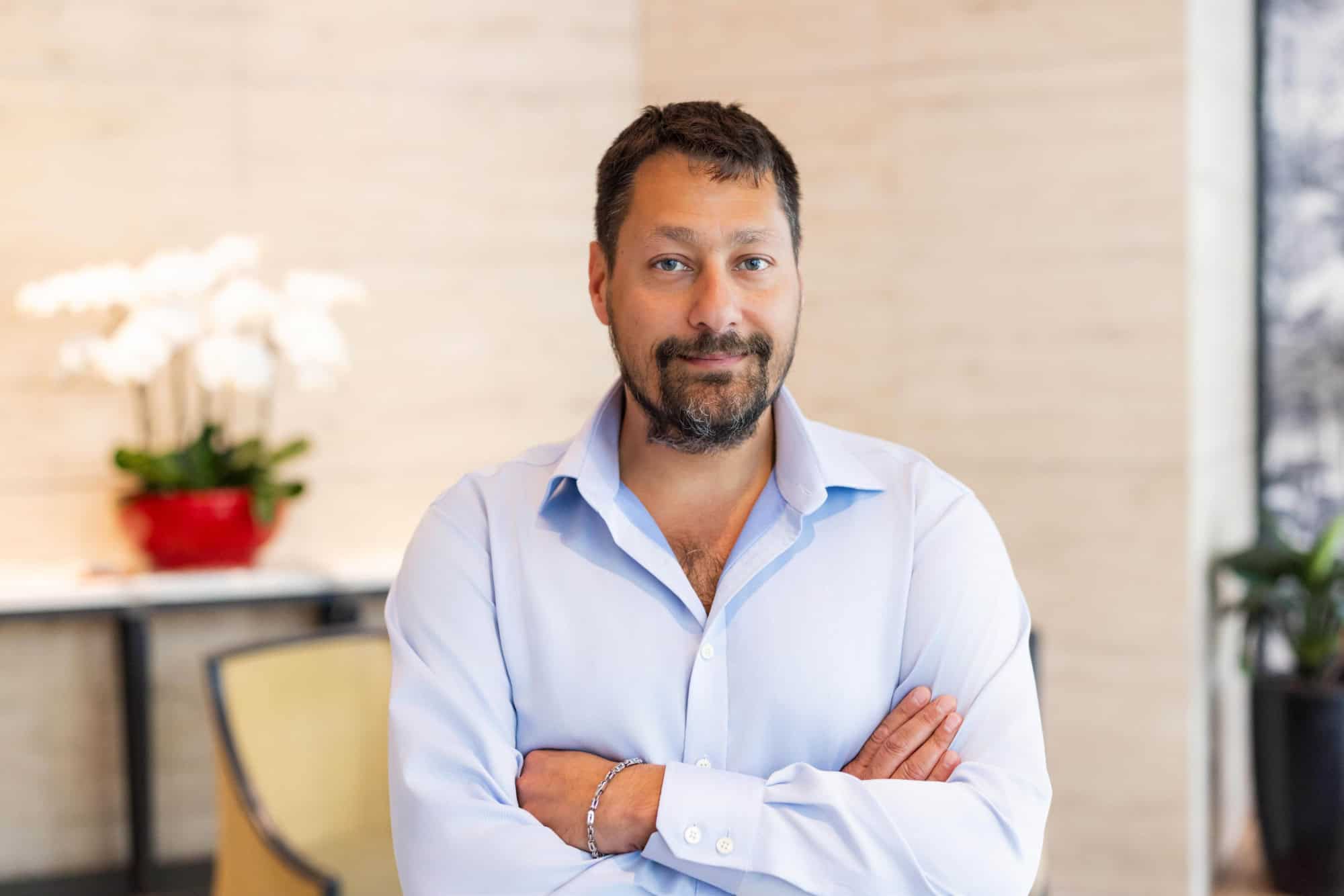 A man with dark hair and beard, wearing a light blue shirt, stands with arms crossed, looking at the camera. A blurred indoor background includes an orchid plant.