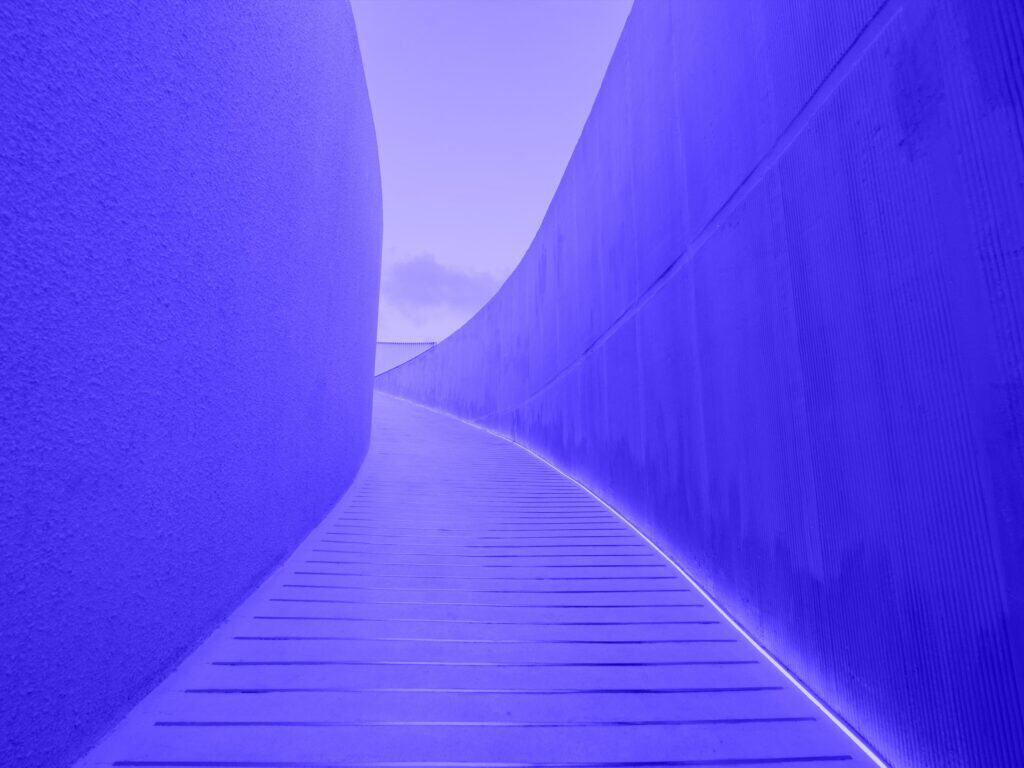 A curved, blue-purple architectural pathway with textured walls and a glowing white light strip along the right wall.