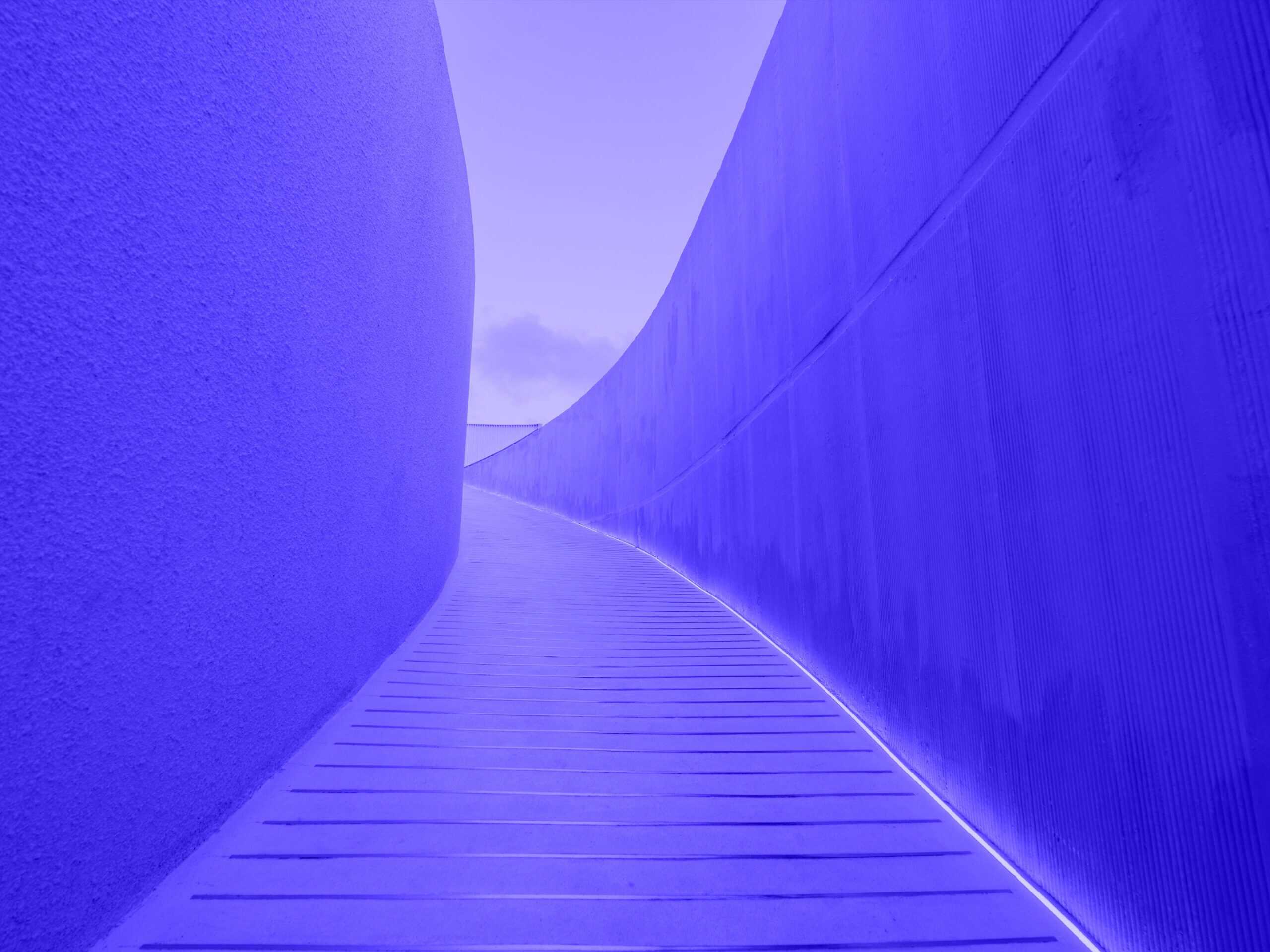 A curved, blue-purple architectural pathway with textured walls and a glowing white light strip along the right wall.